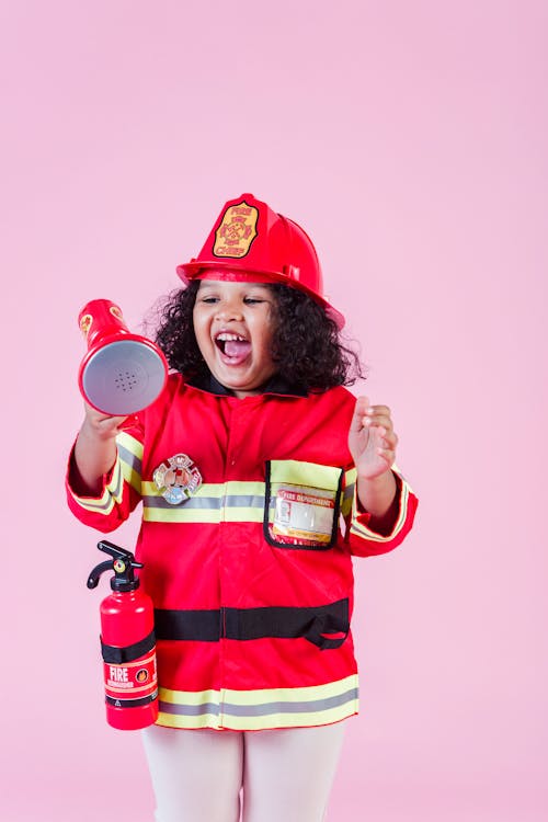 Free Joyful little African American girl wearing bright red firefighter costume and helmet playing with toy loudspeaker in light studio Stock Photo