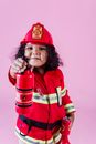 Adorable content little African American girl in bright red firefighter costume pointing toy fire extinguisher at camera while playing in light studio