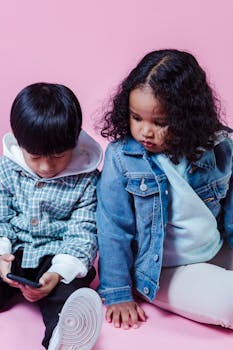 Two children focused on a smartphone against a pink backdrop, highlighting modern childhood.