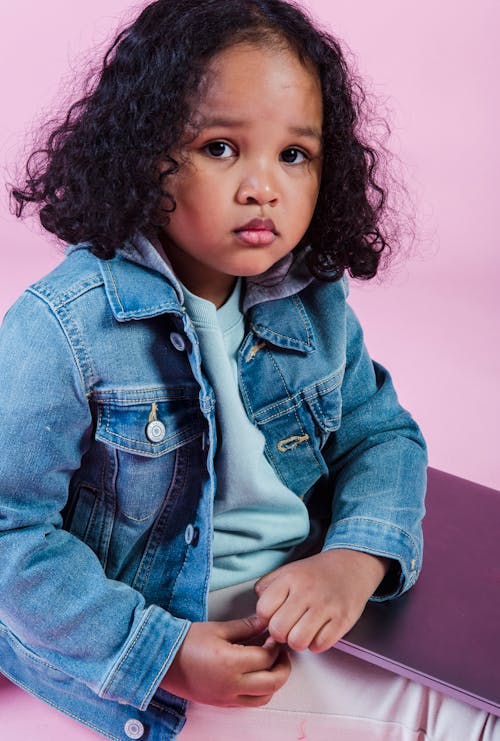 Crop cute calm African American girl in denim jacket sitting on floor with netbook on laps and looking at camera against pink background