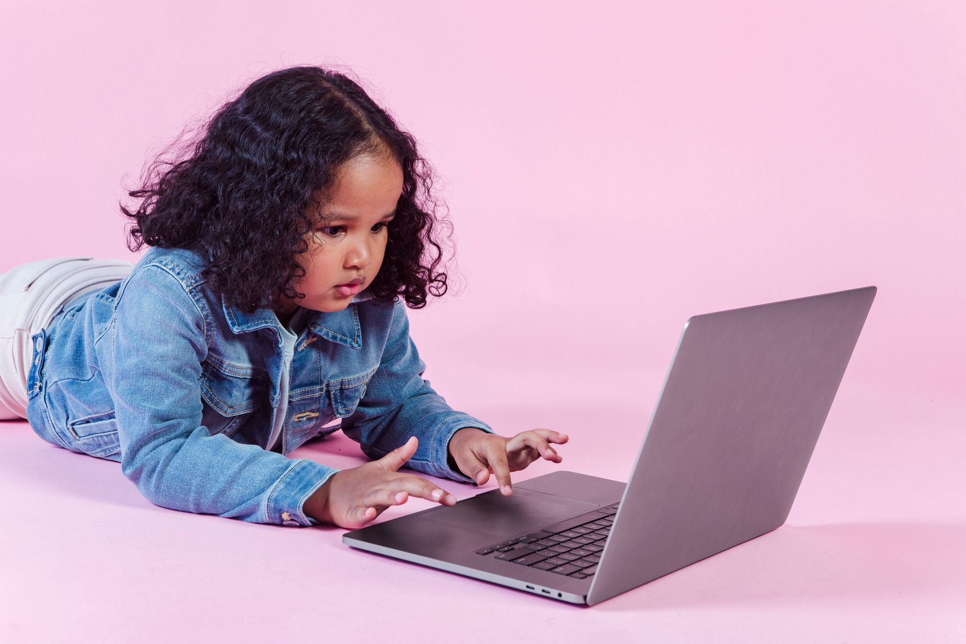 Young girl attentively using a laptop on a pink background, wearing a denim jacket.