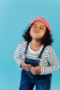 Cute happy African American girl in denim jumpsuit puffing cheeks and grimacing while standing with eyes closed against blue background