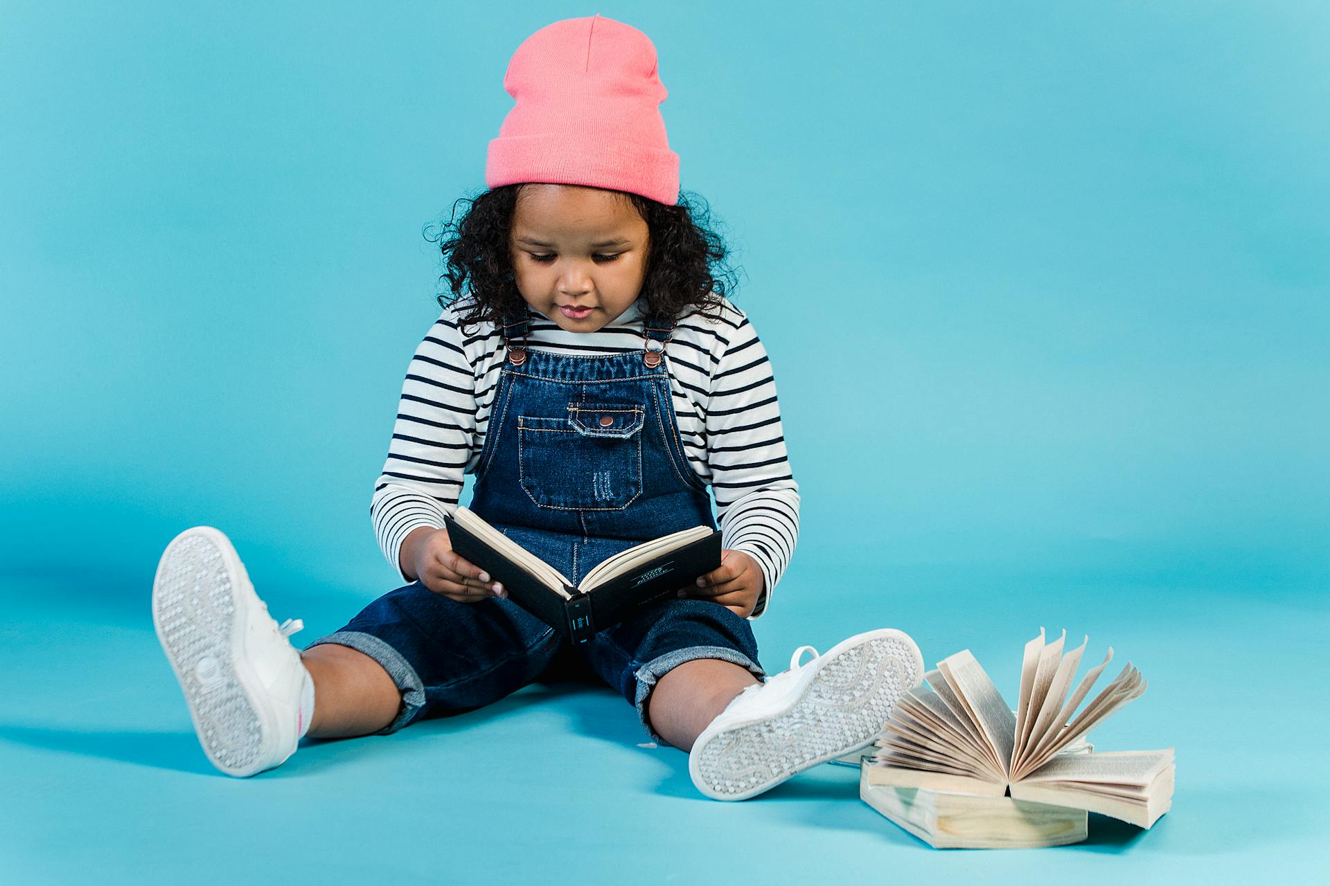 Full body focused little African American girl in trendy outfit reading interesting story and sitting on floor against blue background