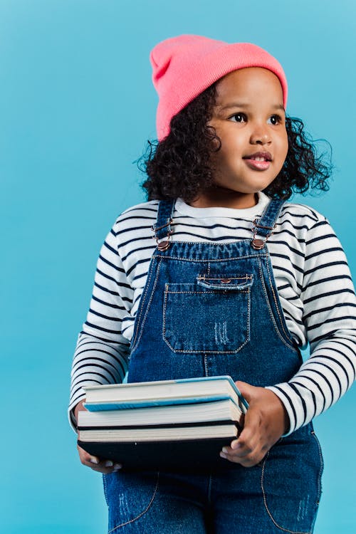 Adorable African American girl wearing trendy outfit and pink hat standing with stacked books in hands and looking away contentedly against blue wall