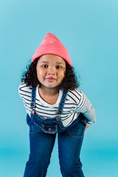 一位活泼的非裔美国小女孩穿着时髦的牛仔连身裤，戴着粉色帽子，在蓝色背景前向镜头弯身