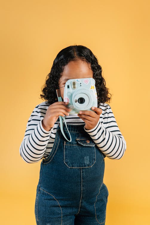 Unrecognizable little stylish ethnic girl in denim jumpsuit taking photo on contemporary instant photo camera while standing against brown background