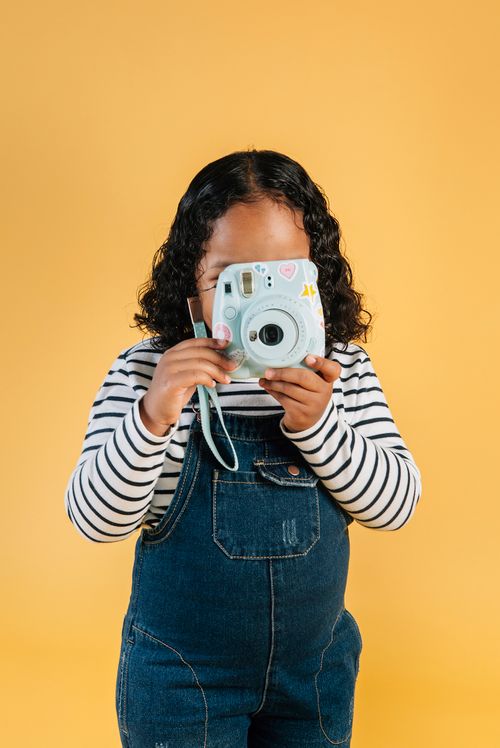 Free Unrecognizable little stylish ethnic girl in denim jumpsuit taking photo on contemporary instant photo camera while standing against brown background Stock Photo