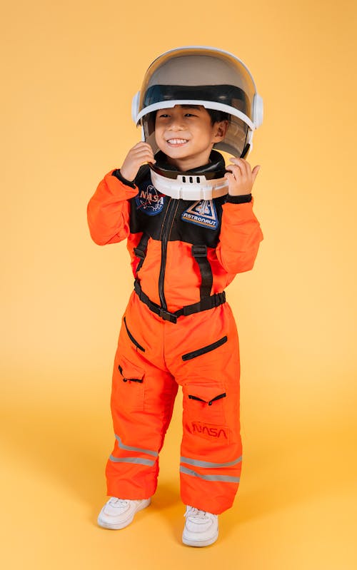 Full body joyful little Asian boy in orange astronaut spacesuit and helmet while standing against brown background and looking away with happy smile
