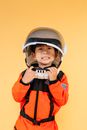 Excited little Asian boy wearing orange astronaut costume touching helmet and looking at camera with toothy smile against brown background in studio
