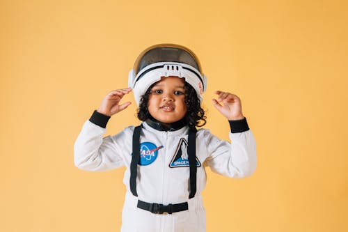 Cheerful little African American girl wearing white astronaut spacesuit and helmet and looking at camera contentedly while standing against brown wall