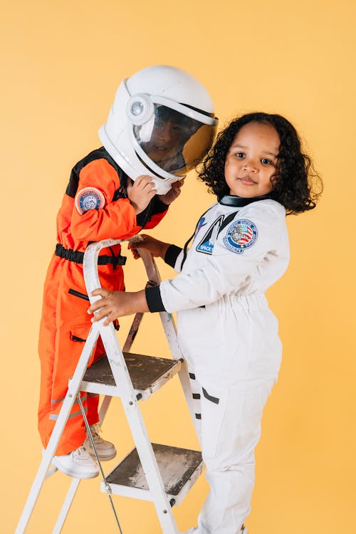 Side view glad little children wearing orange and white cosmonaut spacesuits and standing on stepladder against brown wall in photo studio