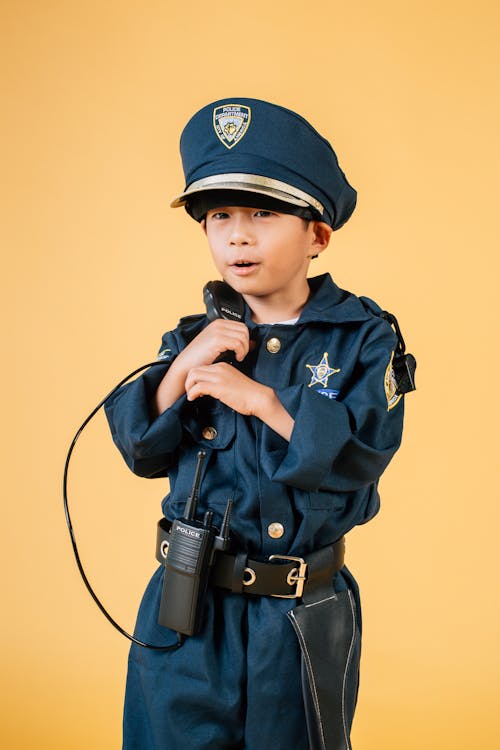 Adorable glad little Asian boy in policeman costume talking on toy radio set while looking away contentedly against brown background