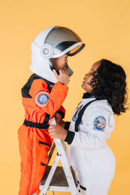 Side view cute multiracial little children in spaceman costumes standing face to face on stepladder while playing together in studio against brown background