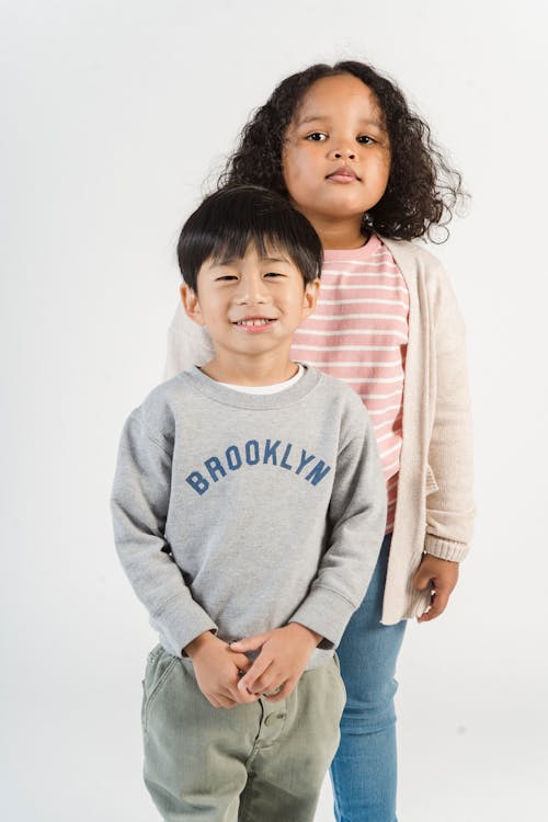 Cute little Asian boy and African American girl in casual clothes standing together against white background and looking at camera