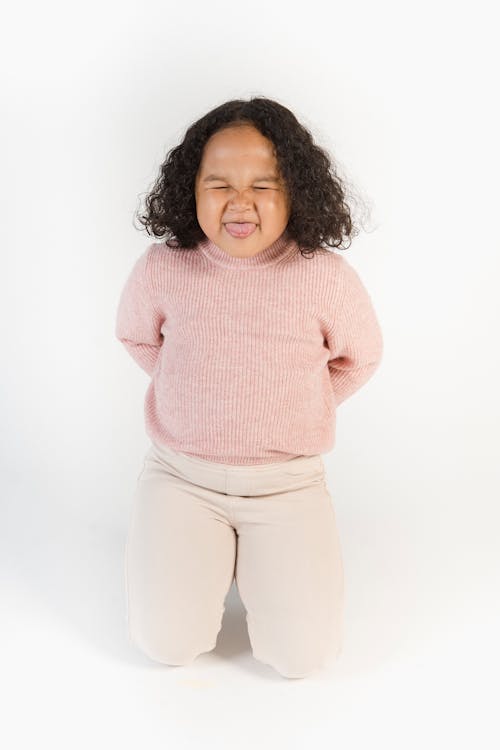 Cheerful adorable little African American girl sitting on knees with hands behind back and showing tongue with closed eyes while playing against white background