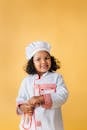 Cheerful little African American girl with curly hair wearing white chefs costume against yellow background and looking at camera