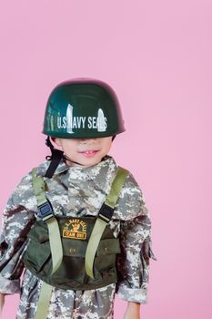Content little boy wearing military costume and helmet lowered on eyes standing against pink wall and smiling
