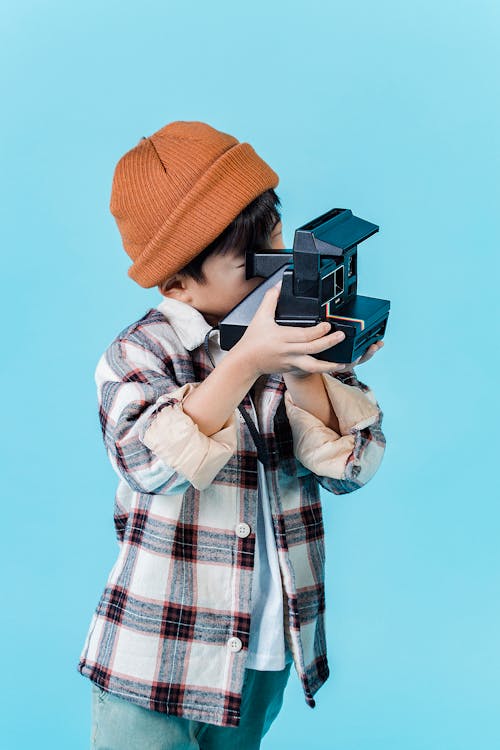 Positive little Asian boy wearing stylish clothes taking photos on retro film photo camera against blue wall in studio