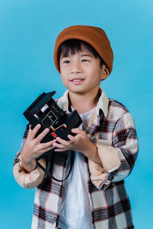 Adorable content Asian boy in trendy outfit holding old fashioned film photo camera and looking away while standing on blue background
