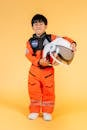 Full length positive little Asian boy in cosmonaut costume biting lip and holding spaceman helmet while standing on yellow background and looking at camera