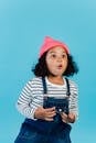 Cute amazed African American girl in cool outfit and pink hat staring away with astonishment while standing on blue background