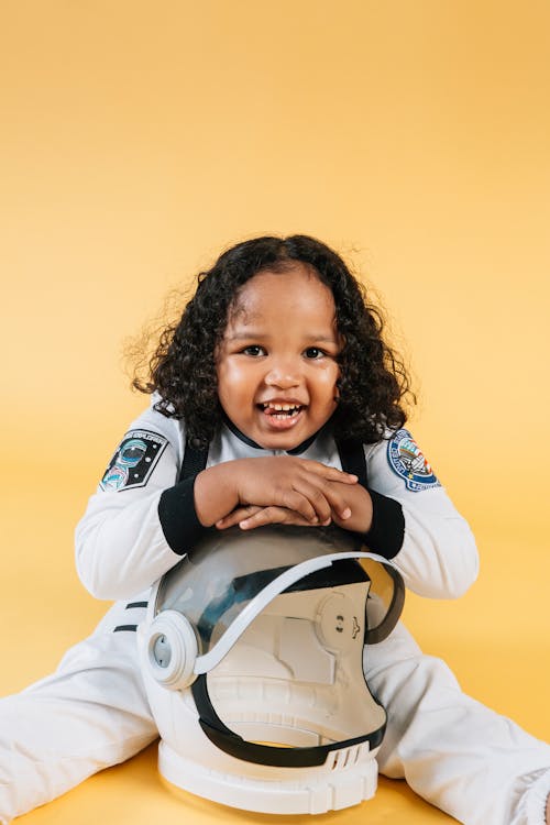 Happy little African American girl wearing astronaut costume and looking at camera with smile while sitting on floor and leaning on helmet against yellow background