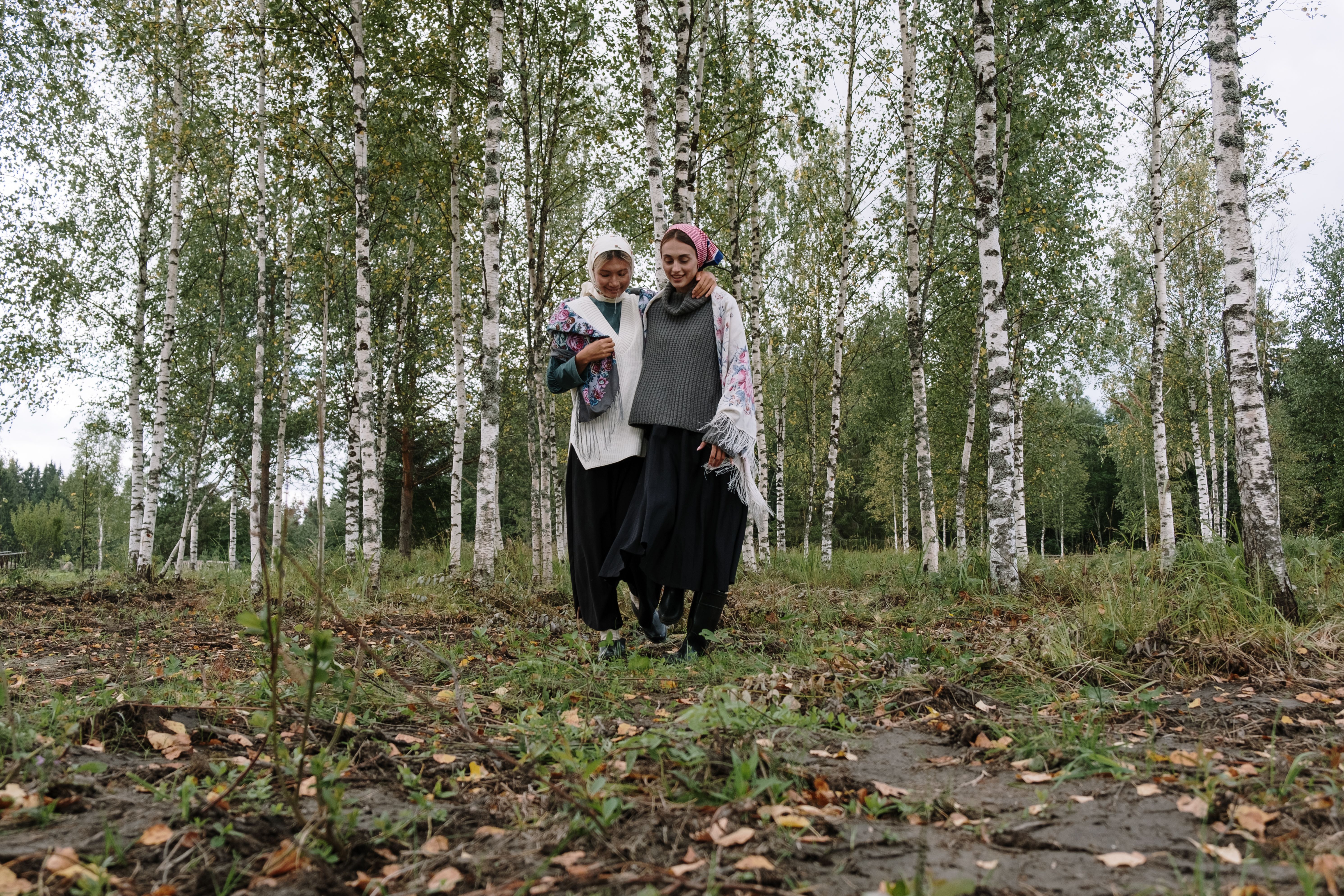 Two women walk with joy amidst a serene birch forest, enveloped by nature's tranquility.
