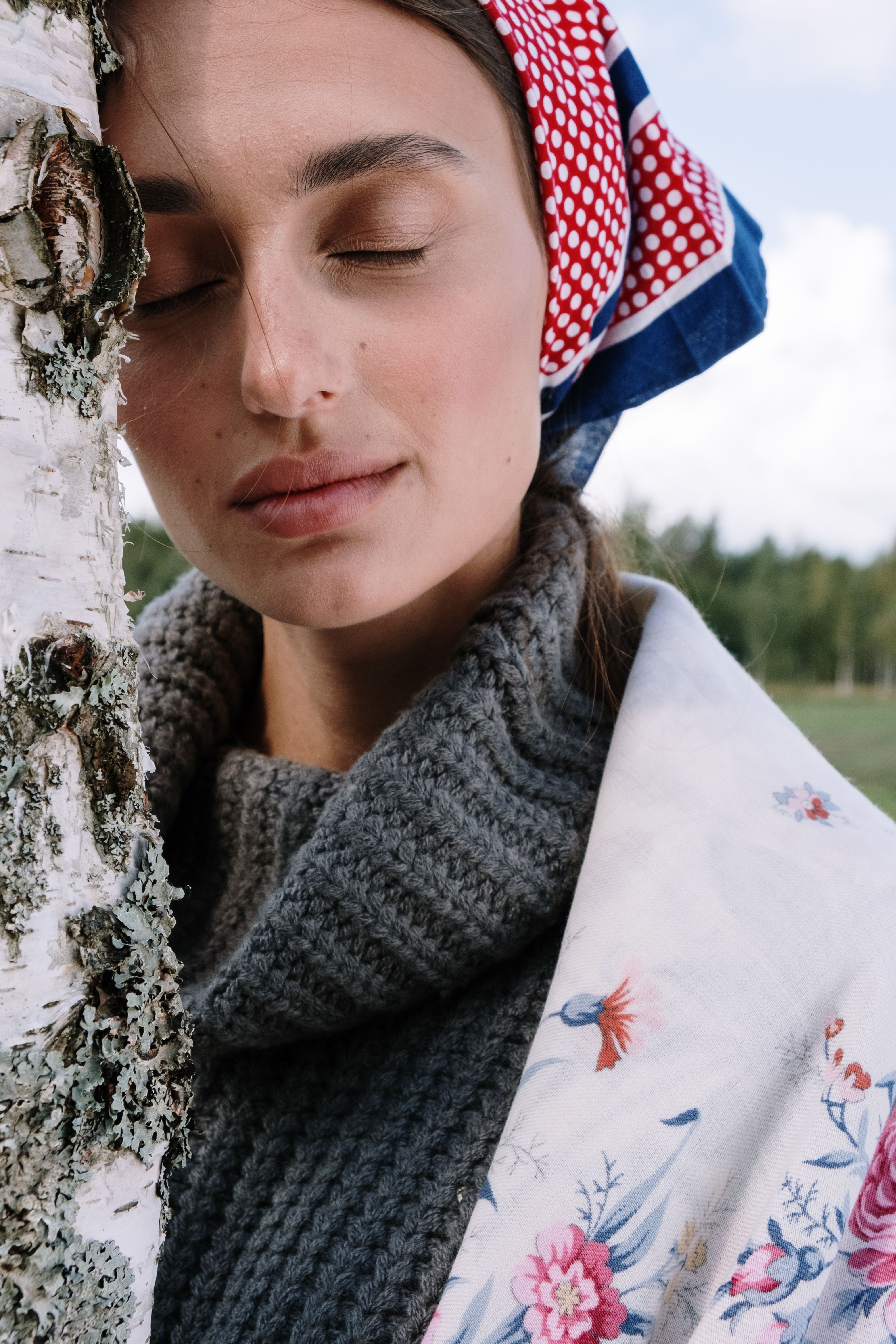 A woman with eyes closed, wearing a scarf, leaning against a tree.
