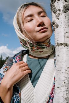 一个闭着眼睛、头戴花卉头巾的女人，静静地倚靠在一棵桦树旁