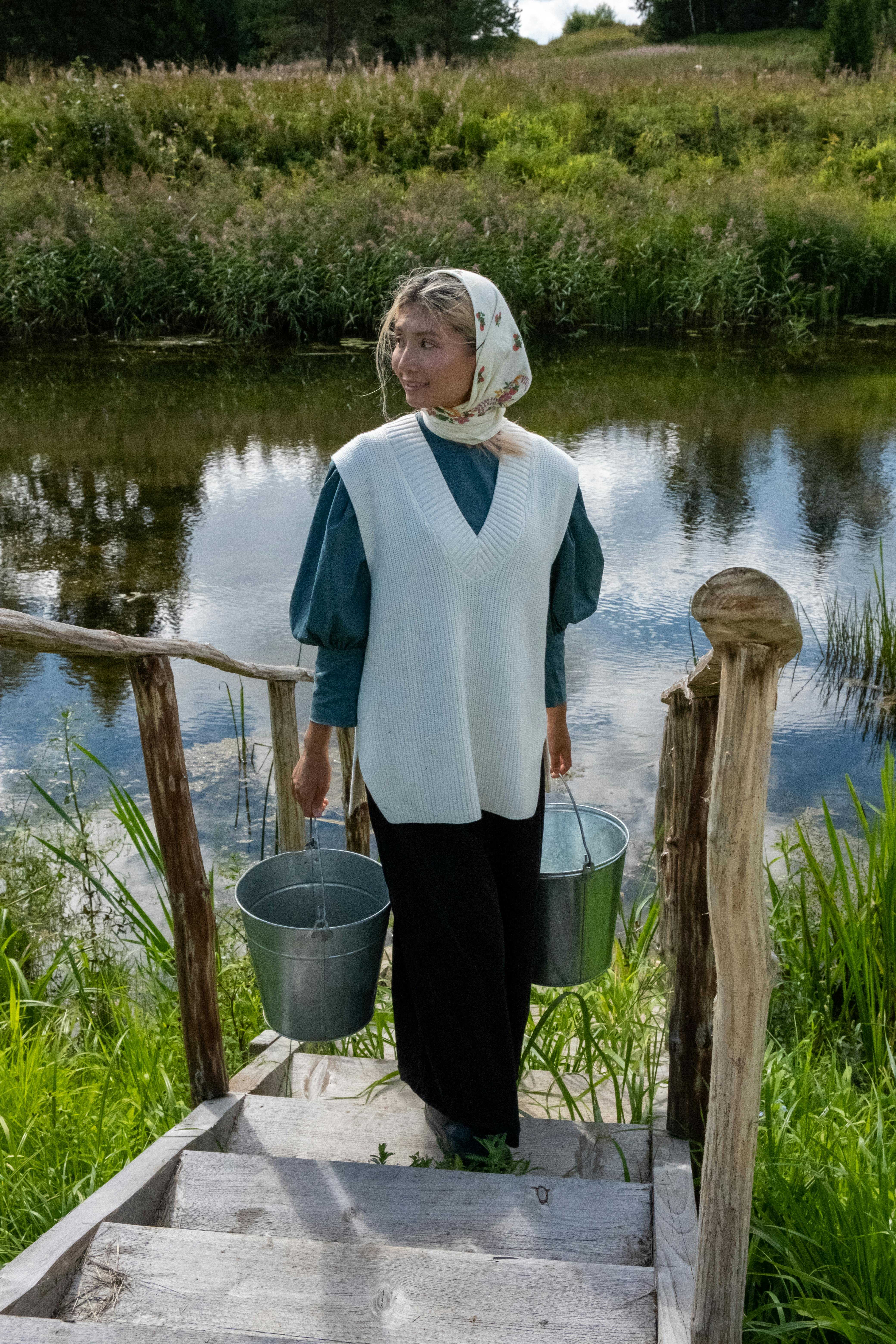 Woman in a headscarf carrying buckets by a countryside pond.