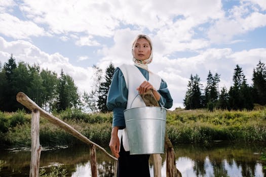 一位年轻女子身着传统服装，站在乡村水道旁，手里拿着一个水桶