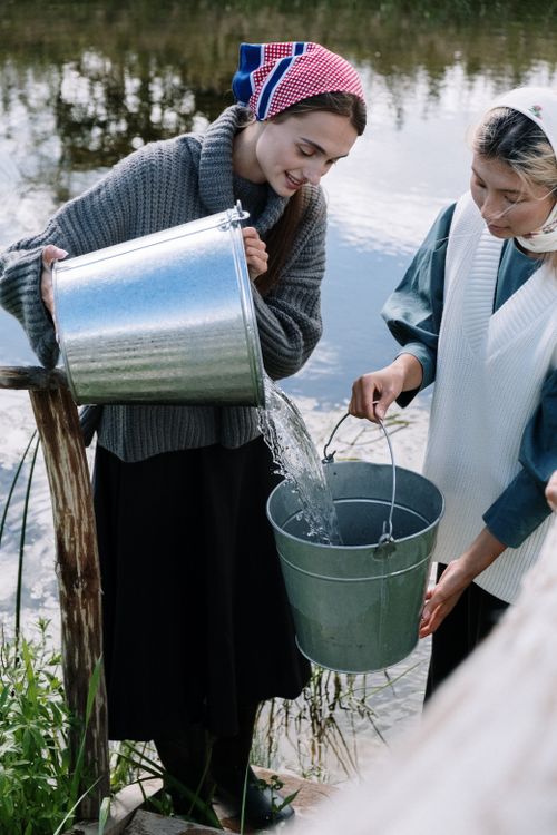 Free Two women in headscarves pour water from buckets beside a serene rural lake. Stock Photo