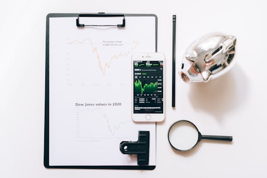 Top view of financial analysis tools including a cellphone, clipboard, magnifying glass, and piggy bank on a white desk.