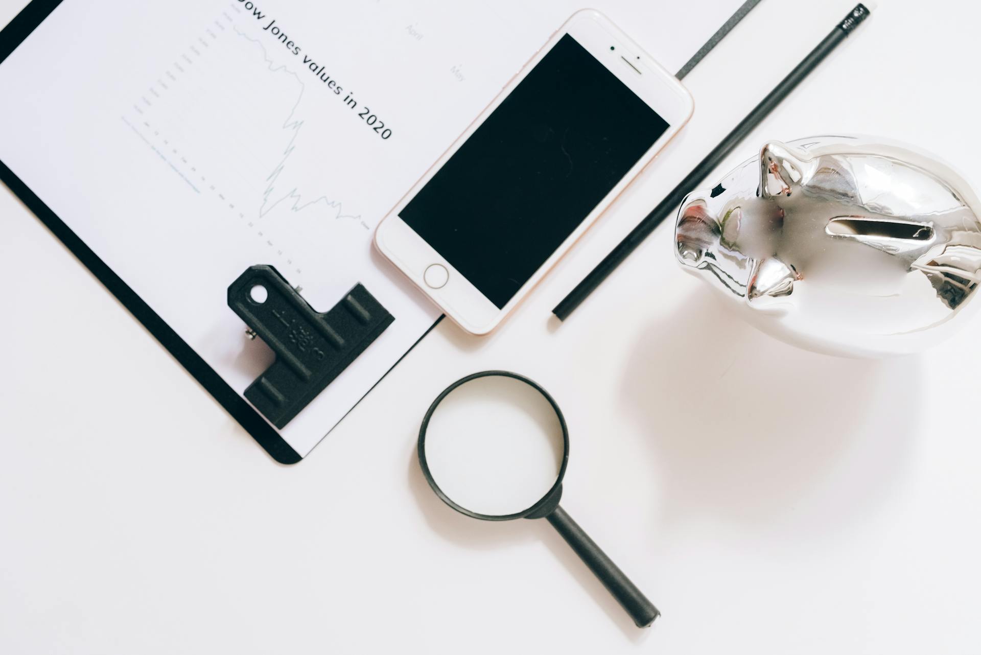 A Smartphone, Documents, Piggy Bank and Magnifying Glass Lying on White Surface