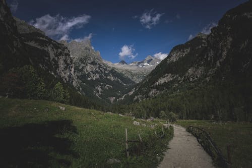 Picturesque scenery of lonely sandy pathway between green grass and rough rocky mountains under blue sky