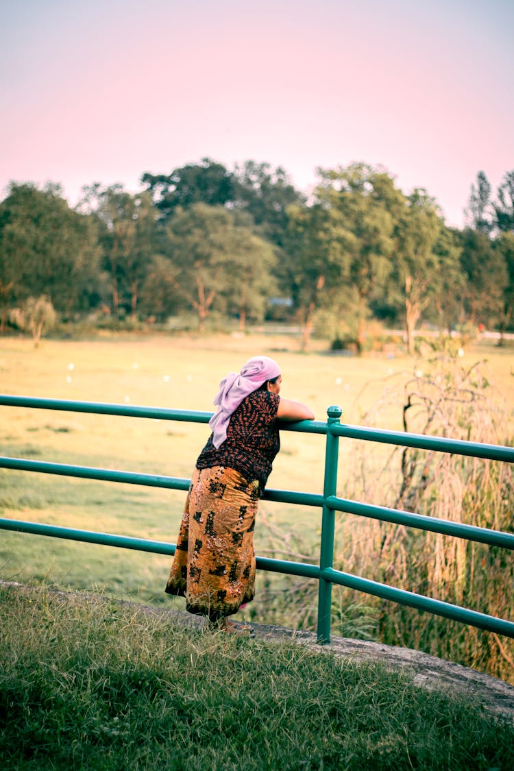 Elderly Woman Resting Against A Fence And Looking At A Field 