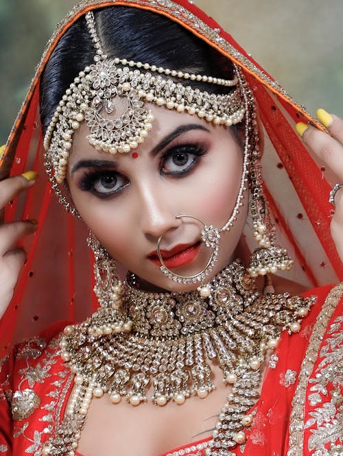 Serious young Indian female wearing red and golden traditional clothes with makeup and piercing in nose with necklace and big earrings with veil on head while looking at camera