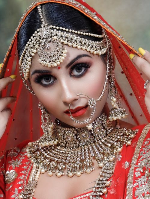 Free Serious young Indian female wearing red and golden traditional clothes with makeup and piercing in nose with necklace and big earrings with veil on head while looking at camera Stock Photo