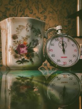 A vintage ceramic cup with floral design and a stopwatch reflected on a surface, capturing elegance and time.