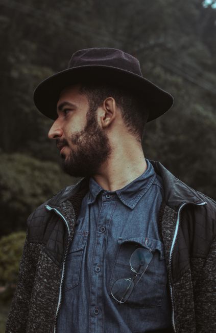 Crop faceless bearded guy in trendy outfit and black hat with glasses near rocky hills and looking away pensively in daytime