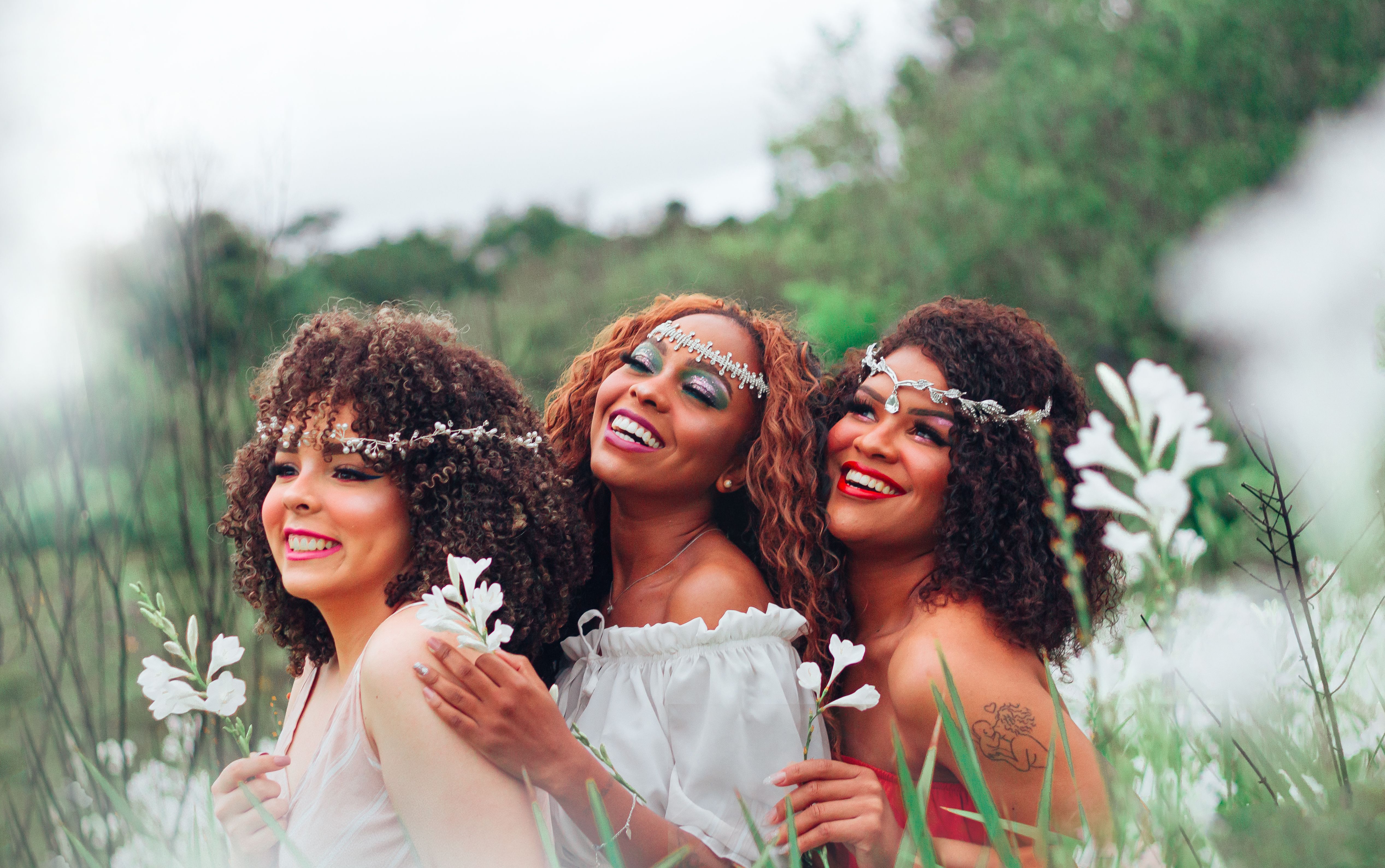 Smiling multiracial female girlfriends with curly hair and wreaths on heads standing in grassy meadow with flowers in hands in summer