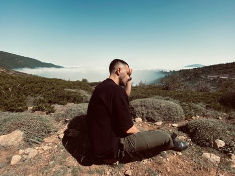 A man sits thoughtfully in a mountainous landscape with a distant view and clear sky.