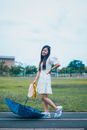 Full body of positive Asian female in dress looking at camera while standing on road with umbrella and streamer pom poms in overcast weather