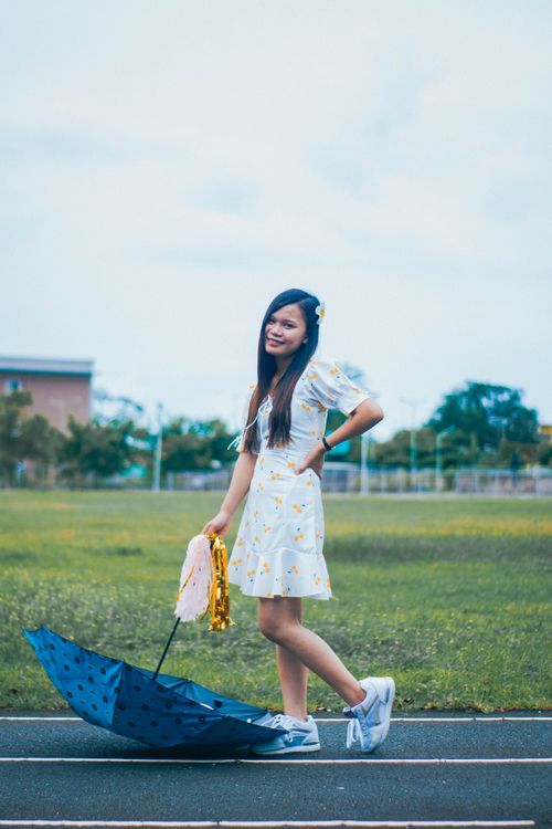 Free Full body of positive Asian female in dress looking at camera while standing on road with umbrella and streamer pom poms in overcast weather Stock Photo