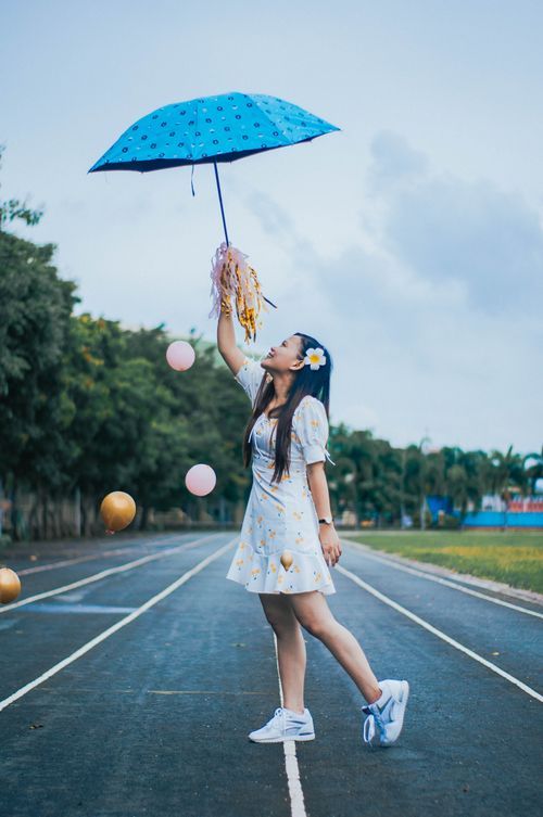 Free Full body side view of positive ethnic female in dress and sneakers standing on running track with umbrella and pom poms among flying balloons Stock Photo