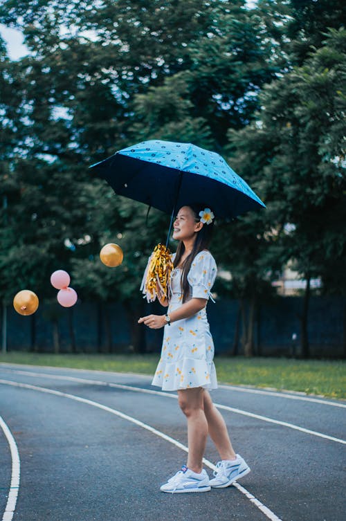 Full body side view of delighted young ethnic female standing with umbrella and pom poms among flying balloons and looking away