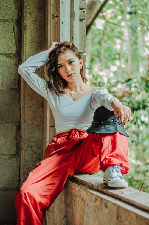 Free Confident young female wearing trendy outfit sitting in window frame of old abandoned building and looking at camera Stock Photo