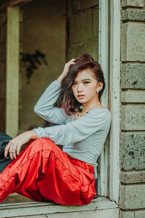 Free Side view of charismatic female in stylish outfit sitting in window frame of old building and looking at camera Stock Photo