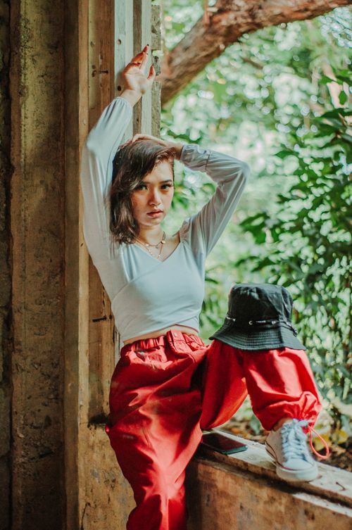 Free Serious Asian female in red pants sitting in window opening near foliage and looking at camera Stock Photo