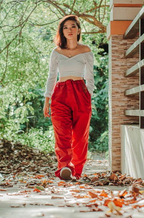 Free Full body of Asian woman in red pants near wall walking on withered foliage against background of green trees Stock Photo